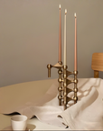 A brass candelabra with three tall taper candles stands on a draped white cloth on a dining table. A white cup is in the lower left, and a wooden chair and bowl are visible in the background against a warm beige wall.