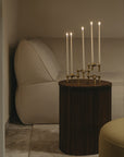 A dark brown fluted side table holding a brass candelabra with five lit white taper candles, set in a cozy living room next to a beige armchair.