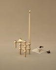 A minimalist brass candlestick holder with a tall taper candle, styled on a warm beige background with small decorative props.