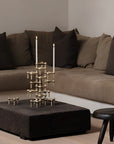 A modern brass candelabra-style candle holder with two lit taper candles sits on a dark textured ottoman in a living room setting with a brown sectional sofa and dark pillows.
