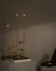 A modern brass candelabra with three tall taper candles lit, displayed on a white pedestal in a dimly lit room next to a dark wooden box.
