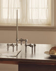 A set of matte gray metal candlesticks displayed on a wooden table in a warm, neutral-toned room with a window and curtains in the background, alongside a plate with a pastry and a ceramic pitcher.