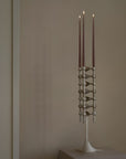 A modern brushed chrome metal candelabra with three taper candles, displayed on a pedestal against a warm beige wall.