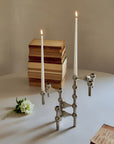 A chrome finished metal candlestick holder with two lit taper candles sits on a white table, styled with stacked vintage books, a small white flower, and an open book against a neutral beige wall.