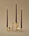 A modern three-stick taper candle holder with a geometric gold metal base, holding three dark brown taper candles with lit flames, set against a warm beige background.