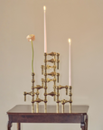 A brass geometric candle holder with three lit taper candles and a single pale pink flower stem, displayed on a dark wooden table against a warm beige wall.