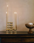 A brass candelabra with three lit taper candles sits on a dark wooden console table against a warm beige wall, with a woven decorative bowl to the right.