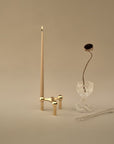 A minimalist tabletop arrangement on a warm beige background: a tall cream candle in a small gold metal holder, a clear glass goblet with a single dried flower stem, and a clear glass spoon.
