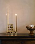 A gold brass candelabra with three lit taper candles displayed on a dark wooden table next to a matching gold pedestal bowl containing white decorative spheres, set against a warm beige plaster wall.