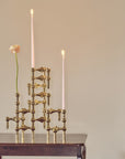 A brass geometric candle holder with three lit taper candles and a single pale pink flower, displayed on a dark wooden table against a warm beige wall.