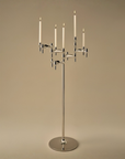 A modern six-arm silver candelabra with white taper candles, set against a solid warm beige background.
