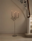 A tall silver floor candelabra with five candle-style lights stands on a staircase landing against a beige wall, next to a white stairwell with a wooden handrail.
