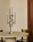 A modern multi-arm metal candelabra with lit taper candles stands on a round light stone dining table, with wooden chairs and a dark wood cabinet in the background.