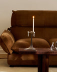 A silver metal candlestick holder with a lit white candle, styled on a dark wood coffee table in front of a brown velvet sofa.