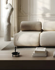 A modern beige sectional sofa in a minimalist, sunlit living room with a light wood coffee table, books, a sculptural white vase and a modern black sculptural bowl cum vase