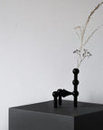 A minimalist black sculptural vase holding a dried grass stem, displayed on a matte black pedestal against a plain light gray wall.