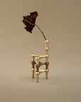 A sculptural gold-toned metal chair with a single dried dark burgundy leaf attached to its back, set against a plain warm beige background.