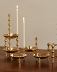 A set of brass candlestick holders and small bowls arranged on a wooden table, with two lit white candles placed among them against a plain beige wall.