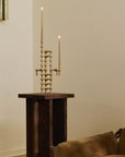 A brass three-arm candelabra with lit taper candles displayed on a dark wooden pedestal in a living room setting next to a brown sofa.
