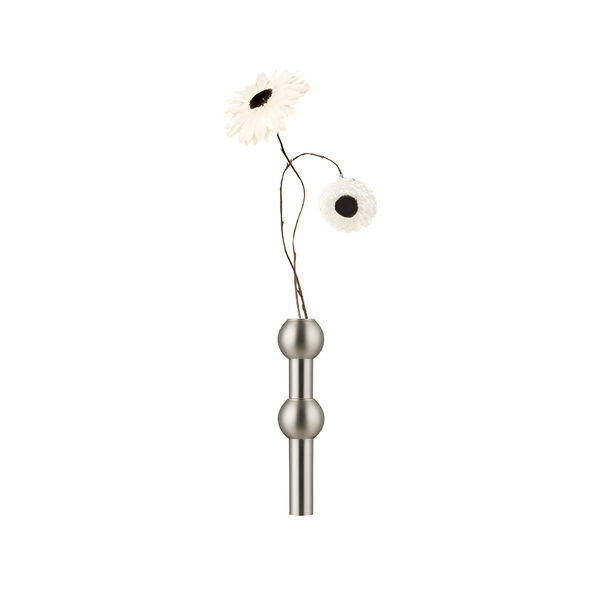 A modern wall-mounted vase with a brushed silver metal finish, featuring two spherical bulb details on a straight stem, holding two white daisy-like flowers with dark centers, set against a solid white background.