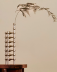 A modern abstract metal sculpture with stacked spherical columns and a dried botanical stem, displayed on a dark wooden pedestal against a warm beige background.