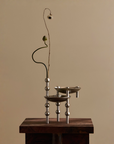 A modern metal flower vase with three tiered bowls and spherical stem supports, styled with a single dried flower stem, displayed on a dark wooden pedestal against a warm beige background.