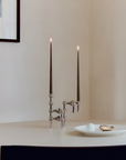 A modern silver metal candelabra with two tall dark taper candles lit, placed on a white table in a minimalist interior setting with a small white plate nearby.