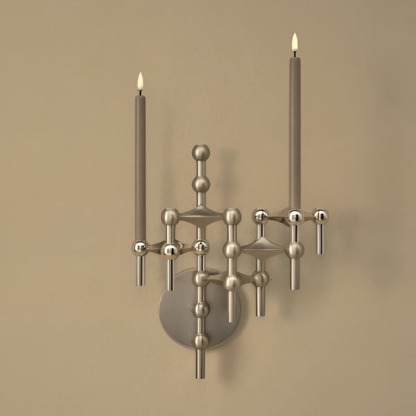 A modern wall-mounted sconce with a brushed silver metal finish, featuring a circular backplate and two tall candle-style arms with lit candles, set against a warm beige wall.