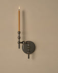A modern wall-mounted candle holder with a matte black circular backplate and articulated arms, holding a single lit taper candle against a warm beige wall.