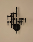 A black metal wall-mounted candelabra with an abstract, geometric branch-like structure and a round backplate, shown against a plain beige wall.