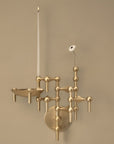 A modern brass wall sconce with a minimalist geometric design, featuring a single candle holder, a tall white taper candle, and a small white flower stem against a warm beige wall.