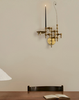 A brass wall-mounted multi-arm candle holder with a single black candle lit, mounted on a beige wall above a wooden table with an open book and a chair.