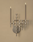 A modern wall-mounted candlelight sconce with a brushed silver metal finish, featuring two vertical candle sleeves with lit flame-shaped bulbs, mounted on a warm beige wall.