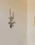 A multi-arm wall sconce with a dark metallic finish and two tall candle-style lights, mounted on a beige wall next to a simpler vertical gold sconce.