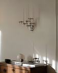 A modern multi-arm wall sconce with a brushed silver finish mounted on a warm beige wall, shown above a wooden console table with books and decorative objects in a sunlit room.