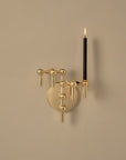 A modern wall-mounted candle holder in a polished gold finish, featuring a circular backplate and three curved arms with spherical accents. One arm holds a lit black taper candle, casting a warm glow against a plain beige wall.