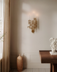 A modern wall-mounted gold sconce with multiple candle-style arms is lit in a styled interior room. The scene includes a cream wall, window with sheer curtains, a wooden table, a white sculptural bust, a tall vase, and a smaller decorative vase on the floor.