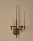 A three-arm wall sconce with a dark bronze metal finish, holding three lit taper candles against a plain beige wall.