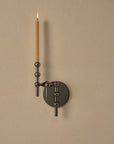A modern wall-mounted candle holder with a matte black circular backplate and vertical support arms, holding a single lit taper candle against a warm beige wall.
