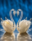 Two clear crystal swan figurines with cream-colored bodies and gold veining, arranged side by side on a reflective surface against a textured blue background.