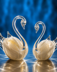 Two crystal glass swan figurines with gold-leaf detailing on their bodies, set against a vibrant blue background with a reflective surface below.