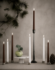 Six led taper candles in white and dark brown shades arranged on a tabletop with silver and black holders, alongside a small green bowl, a pinecone, and a textured neutral wall backdrop with a pine branch wreath.