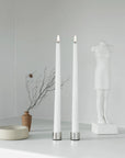 Two tall white led taper candles with silver bases stand on a white surface against a light gray wall, styled beside a small vase with branches, a shallow round dish, and a white mannequin torso.