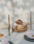 A woven straw basket holding a rustic loaf of bread and a pale pink cloth, styled on a table with two tall matte led taper candles, plates, napkins, and water glasses against a soft marble-like backdrop.
