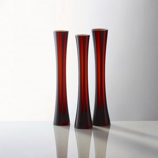 Three tall, slender flared sculptural glass vases in a deep translucent red color, arranged on a reflective white surface against a light gray background.