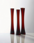 Three tall, slender flared sculptural glass vases in a deep translucent red color, arranged on a reflective white surface against a light gray background.
