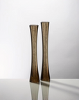 Two tall, slender flared sculptural glass vases in a smoky translucent brown color, standing side by side on a reflective white surface against a light gray background.