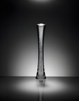 A tall, slender modern sculptural vase with a textured metallic finish, standing on a reflective surface against a dark gradient background.