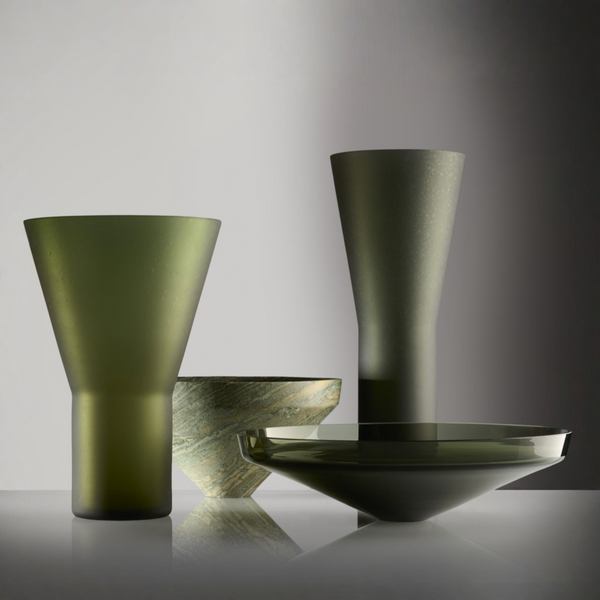 A set of modern glass  art objectsin a dark olive green color, including a conical vase, a tall tapered vase, a shallow bowl, and a wide platter plate, arranged on a reflective surface against a studio background.
