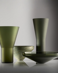 A set of modern glass  art objectsin a dark olive green color, including a conical vase, a tall tapered vase, a shallow bowl, and a wide platter plate, arranged on a reflective surface against a studio background.